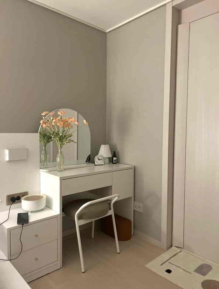 Clean white and gray toned space with a vanity, stool, vase with flowers, clock, and table lamp creating a cozy atmosphere