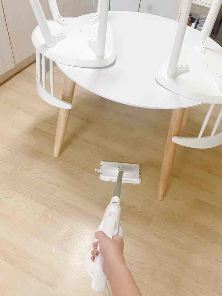 A compact kitchen space featuring a white and wood-tone dining table with chairs, showing a hand performing wet mop cleaning beneath the table