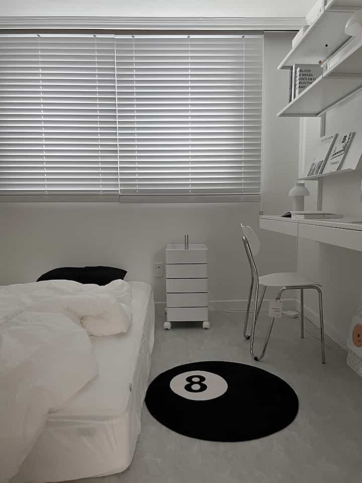 Minimal compact bedroom in clean white tones, featuring a desk and chair on right, and a black round rug with number 8 on center floor