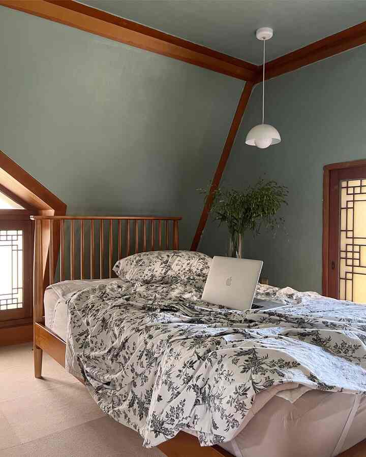 Natural-toned bedroom with wood bed frame and green walls, featuring a plant and white pendant light creating a serene atmosphere