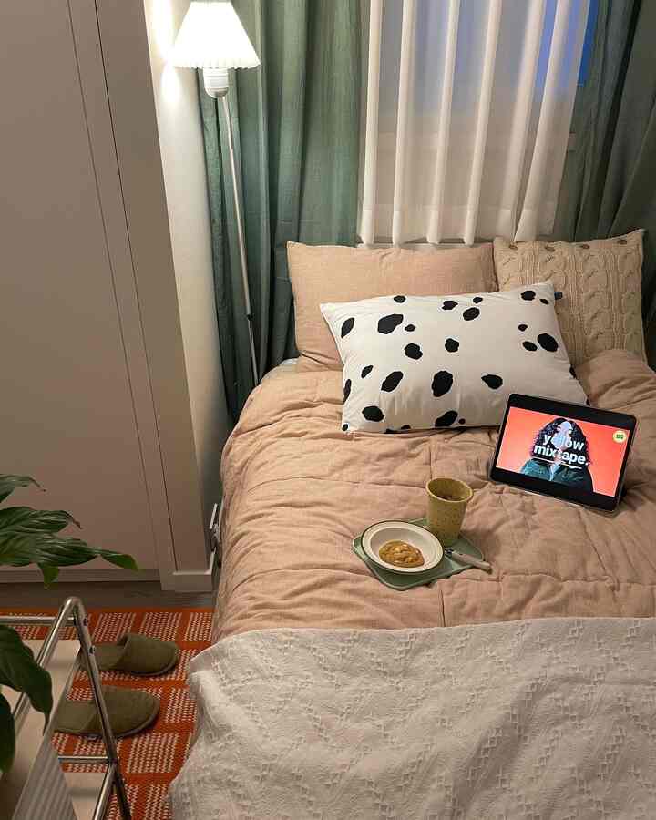 Cozy bedroom with pink bedding and cushions, featuring green curtains and a plant creating a warm atmosphere