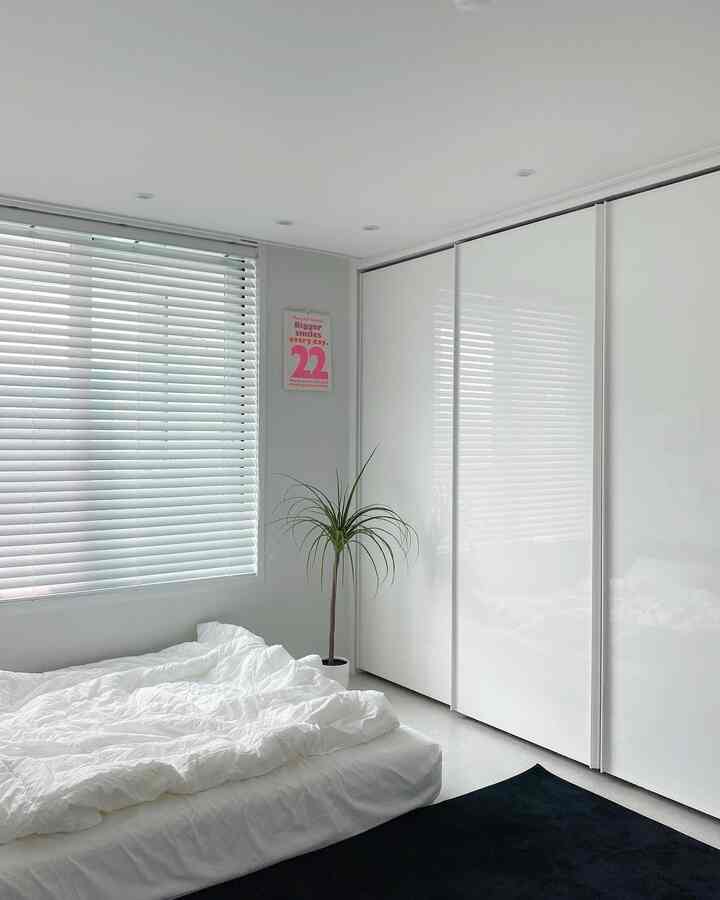 白を基調とした寝室、広い白のスライド式クローゼットとブラインド、白いベッドと自然な観葉植物が配置されたシンプルなインテリア