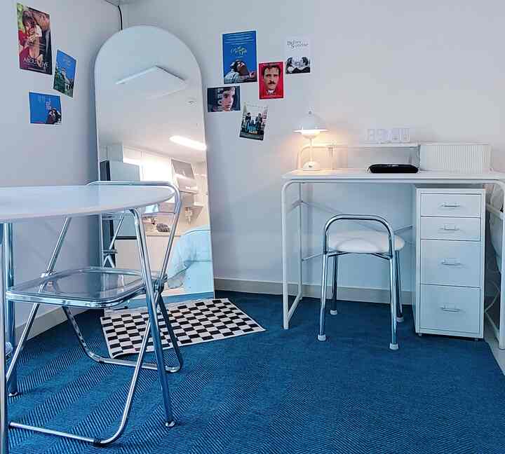 白と青のトーンのワンルーム、白いデスクとクロームチェア、多彩なポスターが配置された印象的な空間