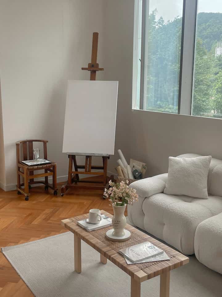 A white and wood-toned workspace featuring an easel, a plush sofa, and a coffee table with a calm, natural atmosphere