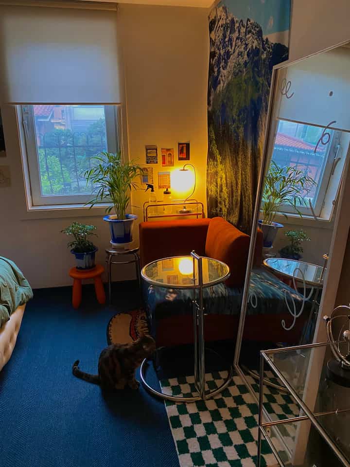 Compact bedroom with warm orange sofa, potted plants, large mirror, and side table creating a modern and cozy atmosphere