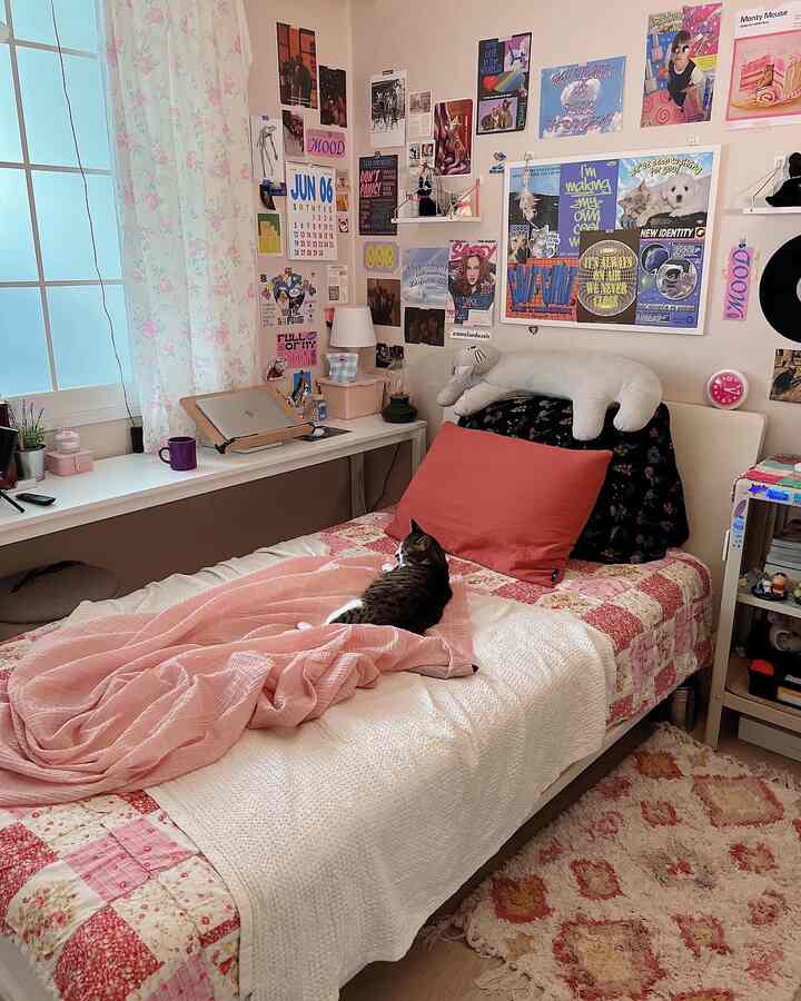 A cozy bedroom in pink and white tones featuring a bed, desk, and walls covered with diverse posters