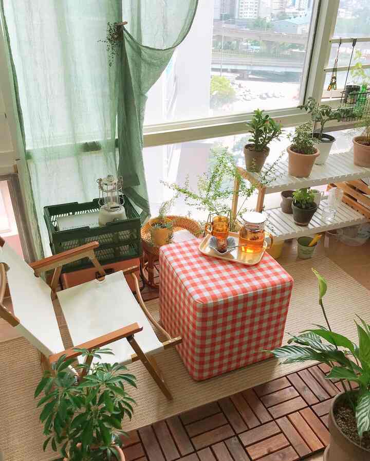 Natural-toned balcony featuring green curtains, various plants, and a checkered stool with a cozy home cafe vibe