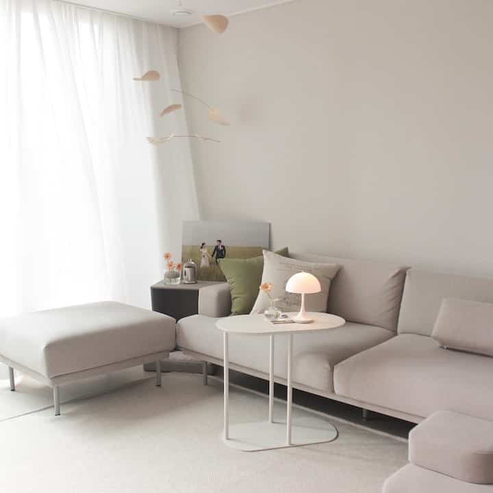 Light beige and white toned living room featuring simple sofa, cushions, side table, and lamp with natural ambiance