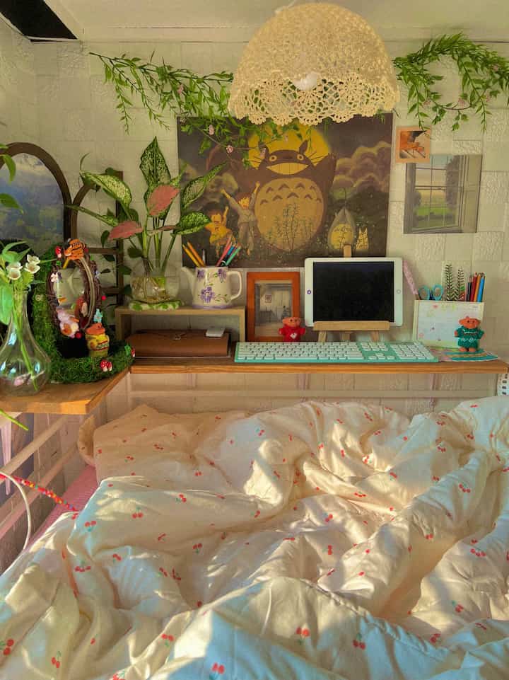A cozy small bedroom with white walls and wood-tone wall shelves, featuring cherry-pattern bedding and a tablet on the desk