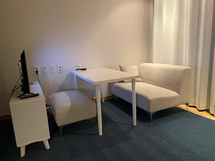 White and blue toned living room featuring a sofa, adjustable leg desk, and blackout curtains in a clean, cozy space