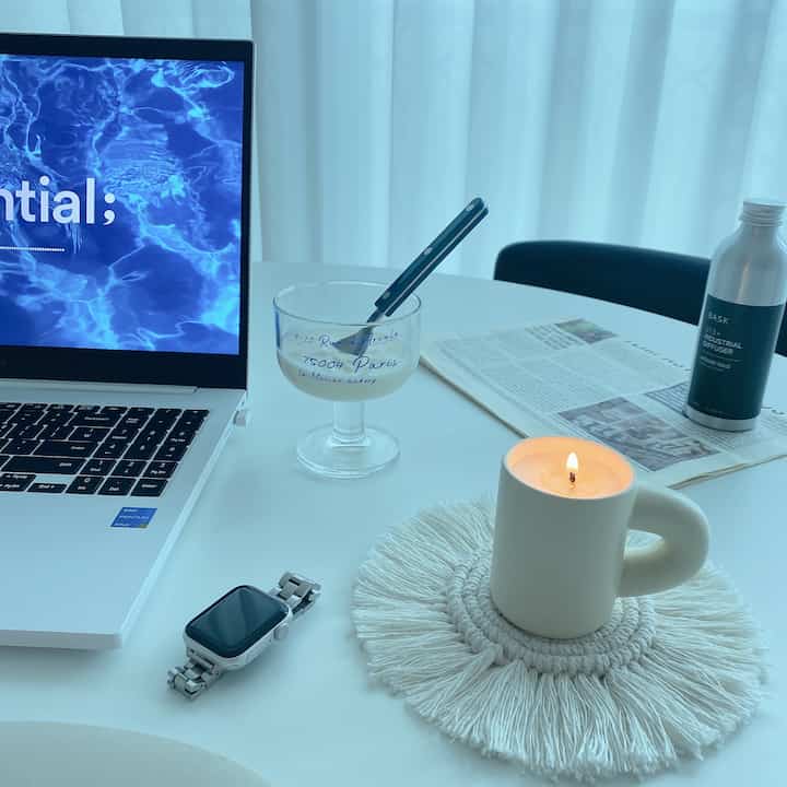 白いカーテンとテーブルがあるホームオフィス空間、ノートパソコン、アップルウォッチ、キャンドル、ディフューザーが配置されたミニマルなインテリア