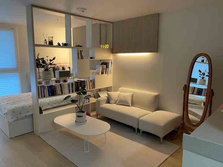 Ivory-toned compact studio living and bedroom space featuring white open bookshelf room divider and wooden framed full-length mirror in a natural modern style