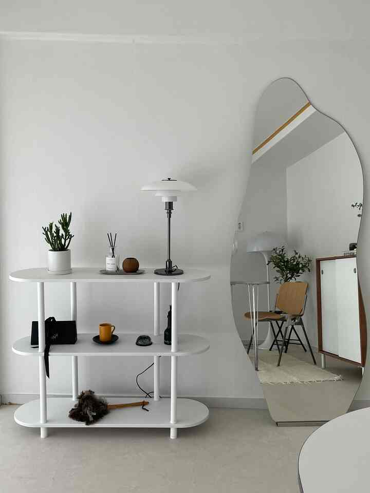 白を基調としたリビングスペース、3段の棚と木目調の椅子、独特な曲線の鏡が配置されたミニマルでモダンなインテリア