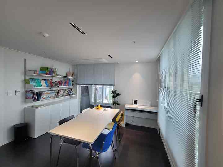 White-toned dining room featuring a long table with colorful chairs, a wall-mounted bookshelf, and aluminum blinds creating a neat atmosphere