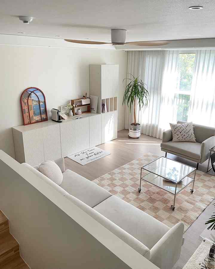 Bright natural-tone living room featuring a large white sofa, glass coffee table, storage cabinets, and a checkered rug with cozy atmosphere