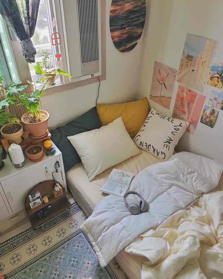 Studio bedroom with white walls and beige bedding, featuring plants by the window, curtains, and posters decorating the wall for a cozy ambiance