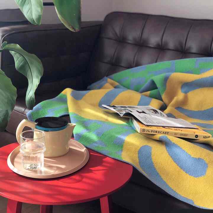 A cozy living room featuring a dark brown sofa draped with a yellow-green blanket, a red coffee table with teapot, bathed in natural sunlight