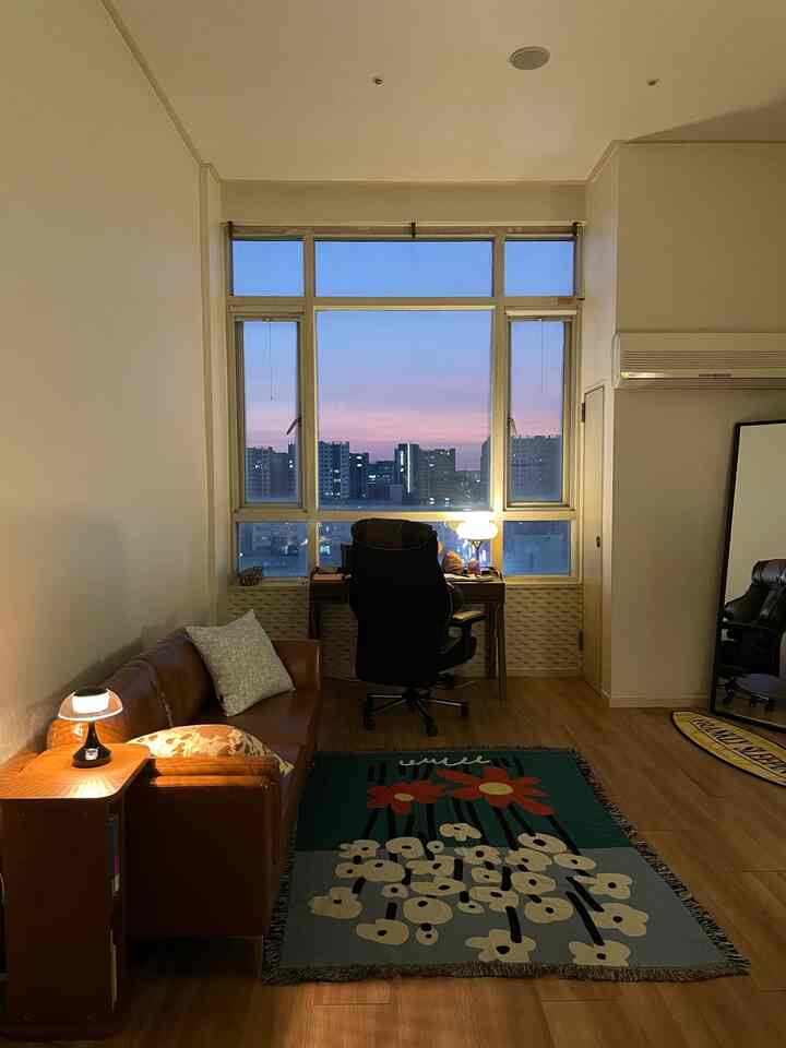 Cozy single household living room and home office with brown and wood tones, featuring a large window overlooking sunset cityscape