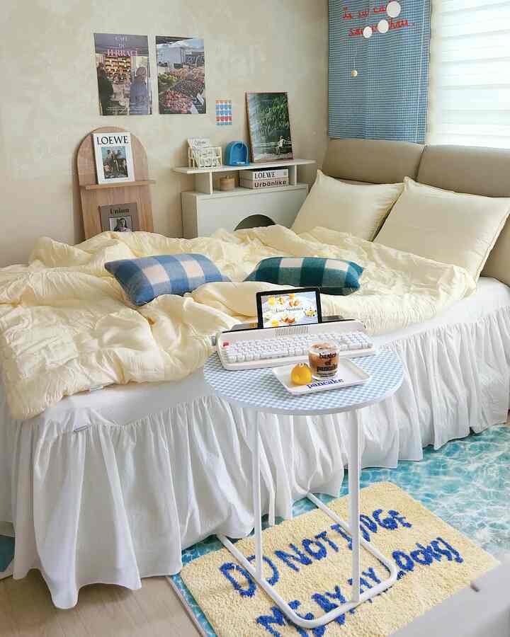 Beige and white toned bedroom featuring a sofa bed with soft yellow duvet and blue checkered cushions, accompanied by a round side table in a natural modern setting