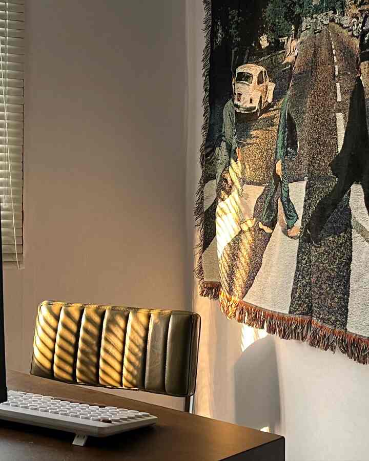 温かみのあるブラウンの木製デスクとオリーブグリーンのパディングチェアがあるホームオフィス空間、右側にはビートルズのタペストリーが壁に掛かっている