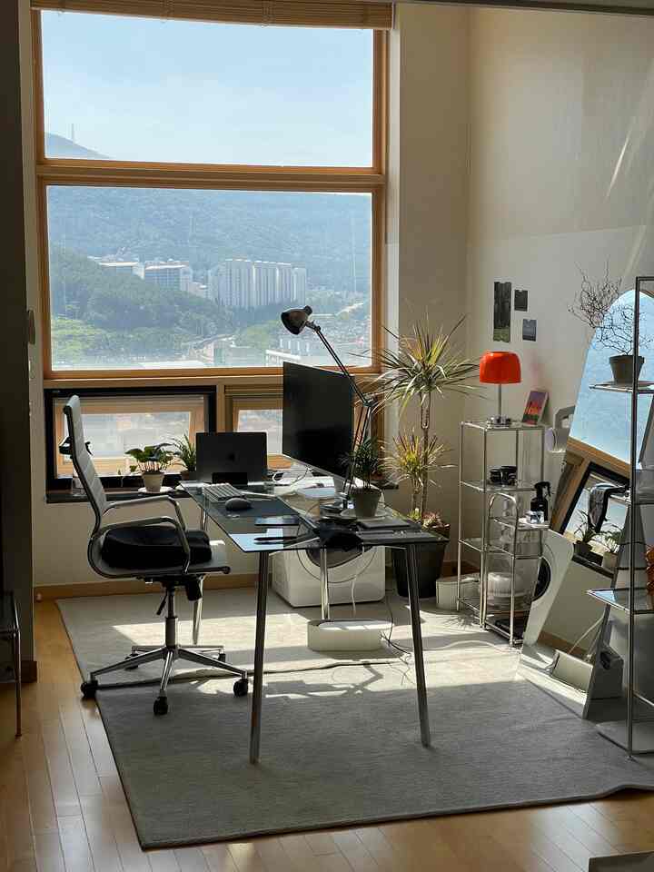 Bright white and beige wood-tone home office featuring a glass desk, metal chair, and various plants in a modern workspace