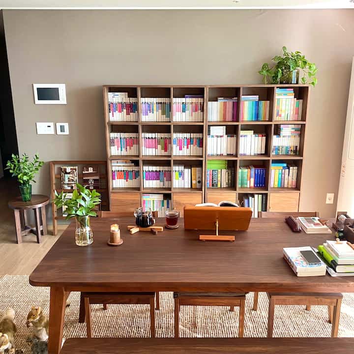 A beige-walled study and living room space featuring warm brown wooden furniture, bookshelves, and plants creating a natural and cozy atmosphere