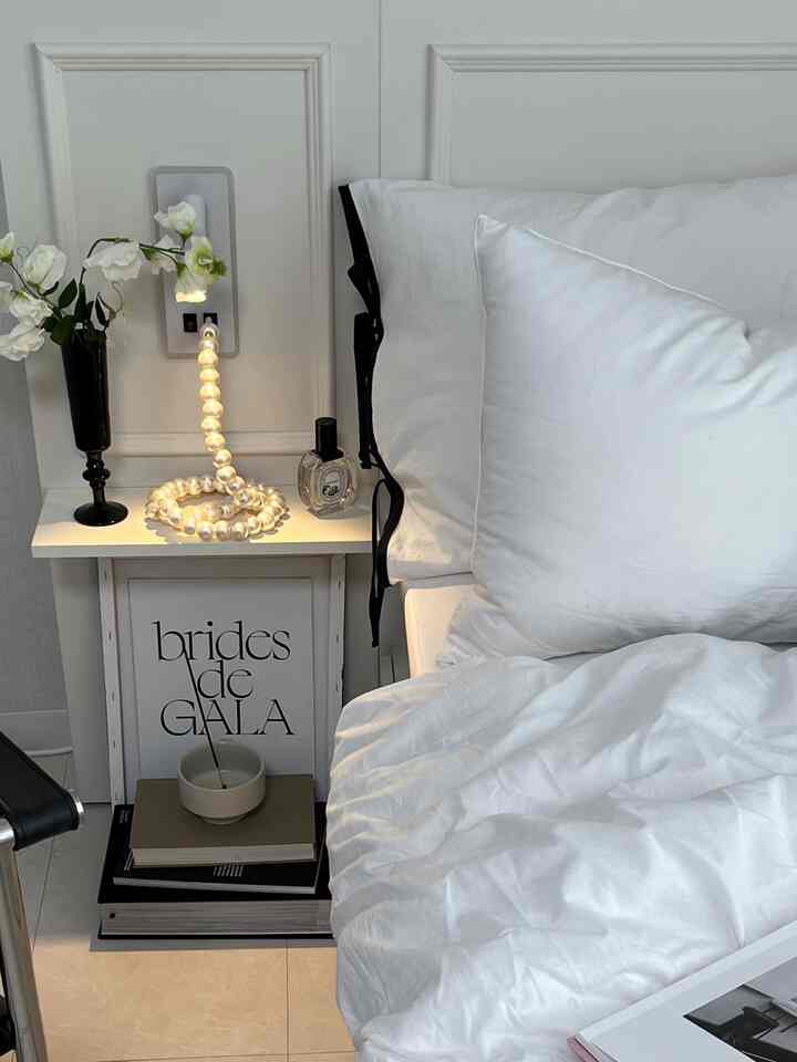 Clean white-toned bedroom featuring white bedding, a nightstand with pearl lamp and vase, in a simple setting