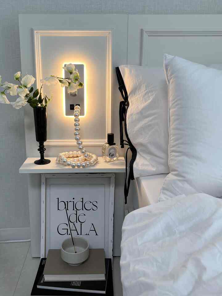 White-toned bedroom nightstand featuring a vase and illuminated wall panel in a minimal setting
