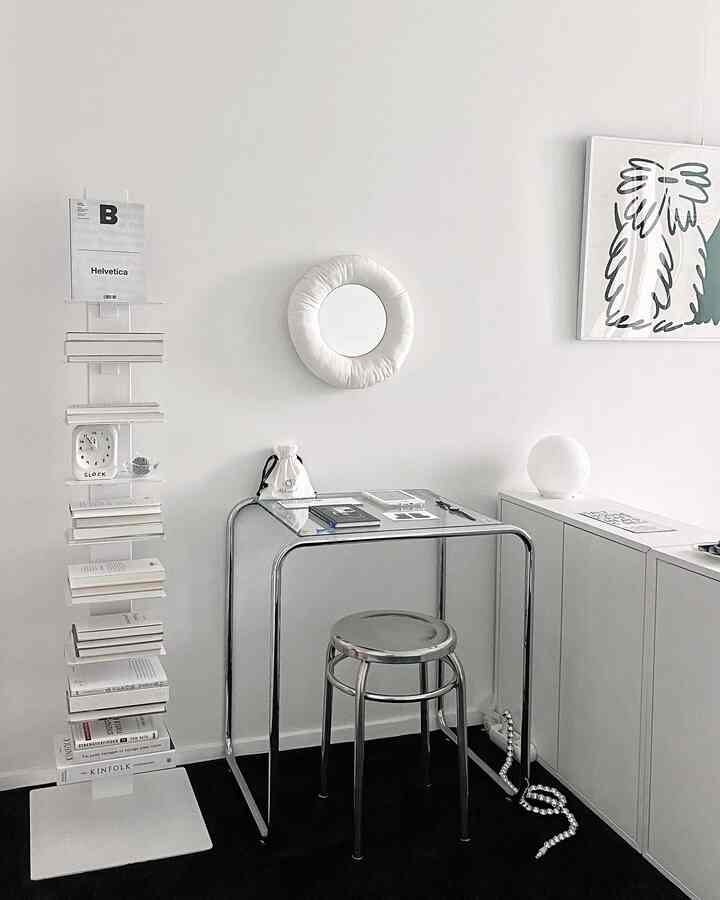 Minimal home office space in white and silver tones featuring tempered glass desk, stainless round stool, and donut-shaped wall mirror with clean decor