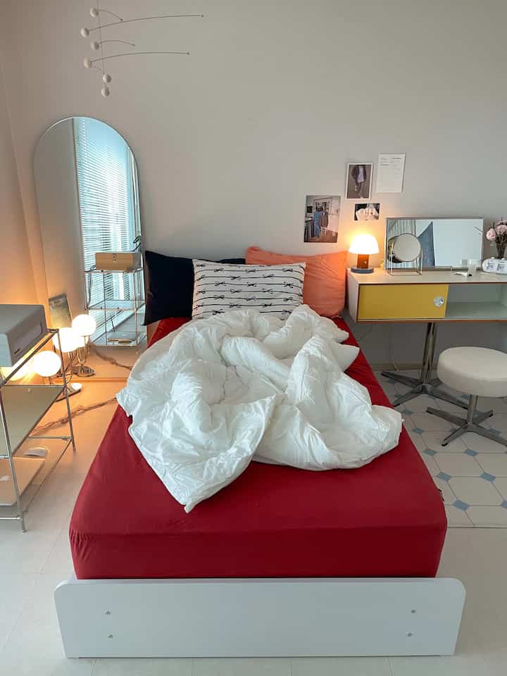 Modern single household bedroom featuring a red mattress, white duvet cover, vanity with stool, and a calm atmosphere