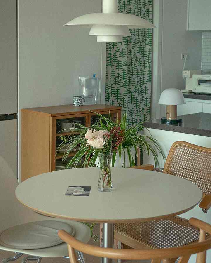 White and wood-tone kitchen and dining room featuring a round dining table, rattan chair, and green plants creating a natural ambiance