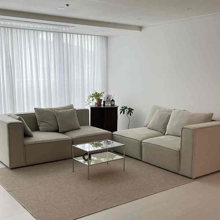 A white and beige living room featuring sectional sofa, glass table, and sheer curtains with a simple modern vibe