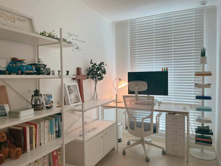 A small study space in white and wood tones featuring a simple desk, bookshelf, and prayer cross, creating a cozy and calm atmosphere