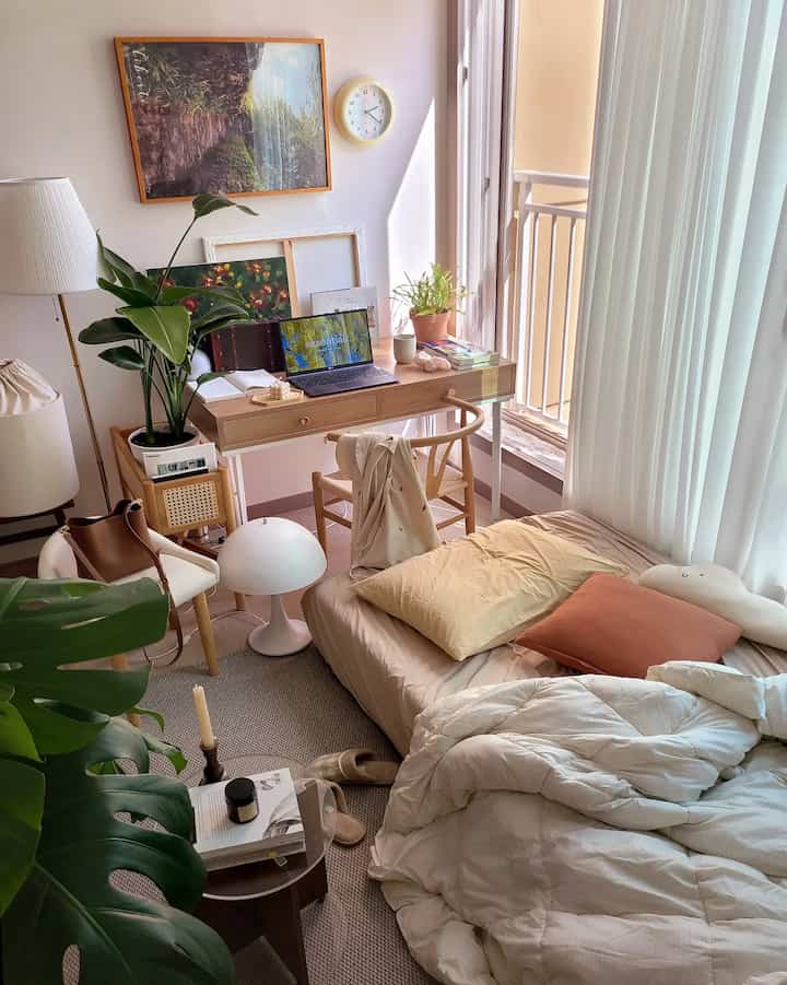 ベージュとホワイトの色調のワンルーム寝室、机と椅子、カーテン、植物が配置された落ち着いた自然なインテリア