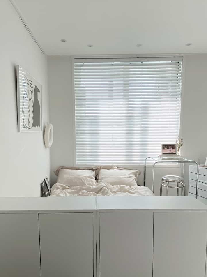 A white-toned small bedroom featuring a beige bed with bedding, transparent stool, and tidy drawer cabinet in a minimal setting