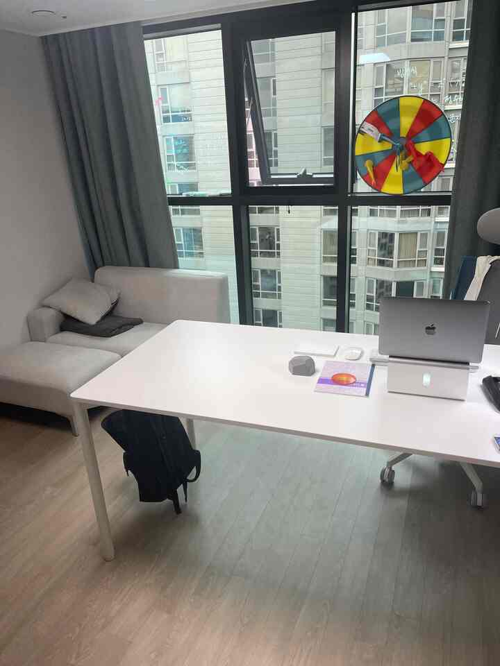 Compact home office in white and gray tones featuring a large central desk and beige sofa on the left, presenting a clean workspace