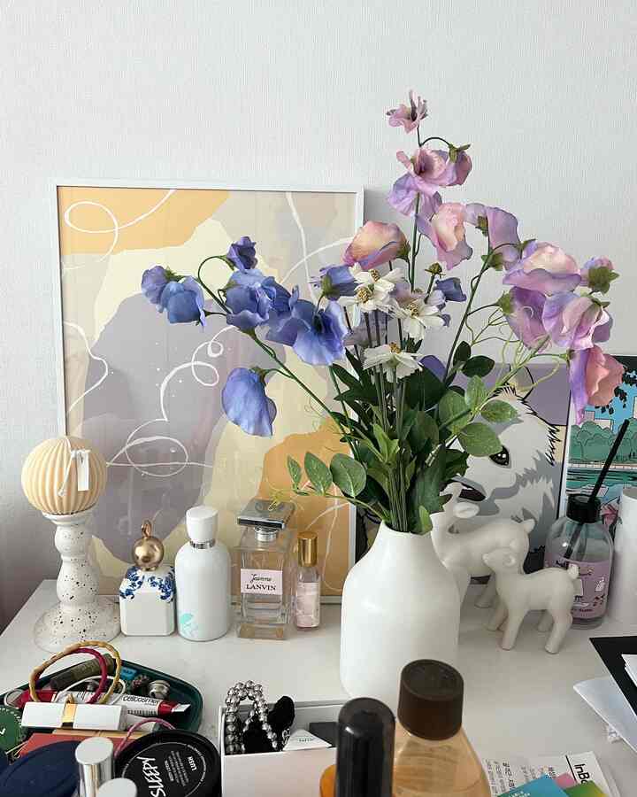 Interior space with white background, a white vase holding pink and blue artificial flowers on a small table with various decorative items
