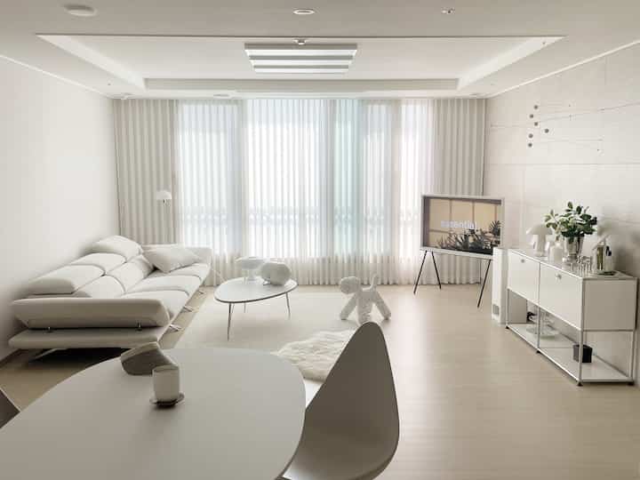 Bright white-toned living room featuring large fabric sofa, sheer curtains, minimalist sideboard and coffee table in a modern setting