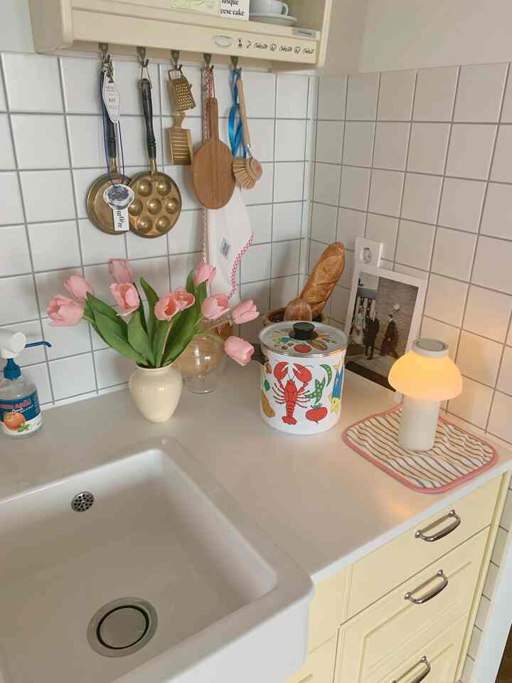 Cozy kitchen with bright yellow cabinets and white tiled walls, featuring a lit table lamp and pink artificial tulips on the countertop