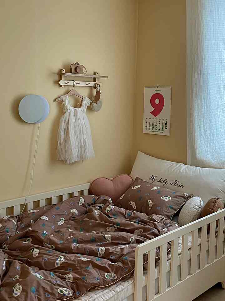Yellow-toned kids' room featuring white toddler bed with brown character bedding, wall shelf, and calendar creating a cozy atmosphere