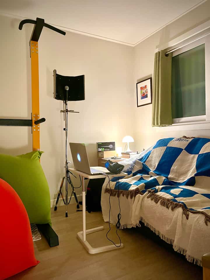 A small home office with white walls and wood tone flooring, featuring a sofa draped in a blue checkered blanket, bean bags, and a table lamp in a cozy mini studio