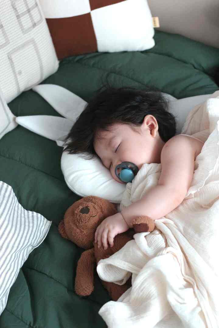 暗い緑色の掛け布団と茶色のぬいぐるみが置かれた子供部屋のベッドで赤ちゃんが穏やかに眠る印象的な空間