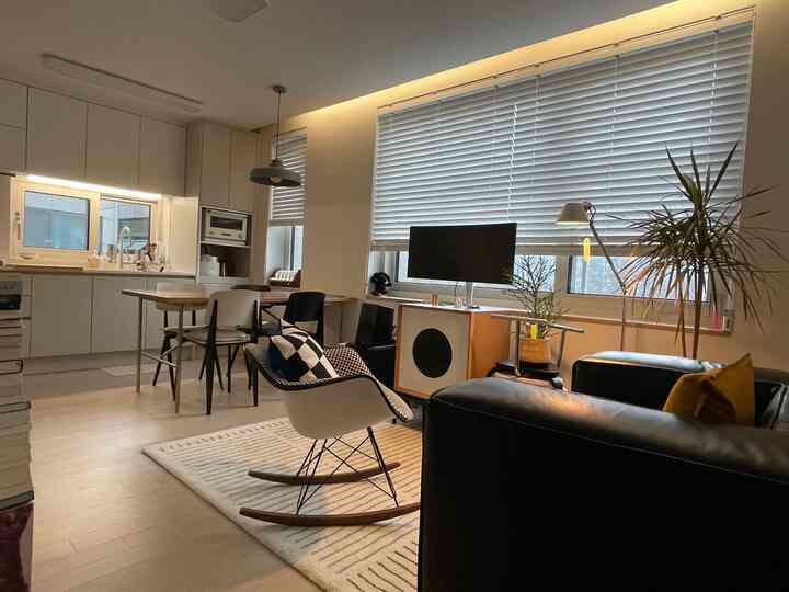A softly lit natural-toned living room and kitchen featuring an Eames armchair and blinds with modern decor