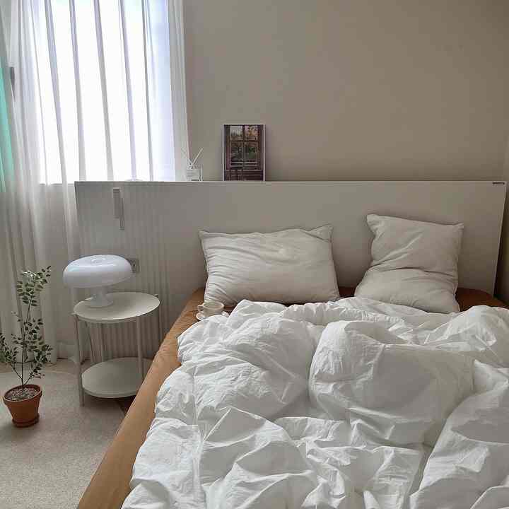 Natural-toned bedroom space featuring white mattress and pillows, brown mattress base, bedside table, and plants creating a cozy atmosphere