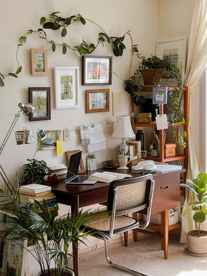 Natural beige-toned workspace featuring wooden furniture, rattan chair, multiple picture frames, and plants creating a cozy atmosphere
