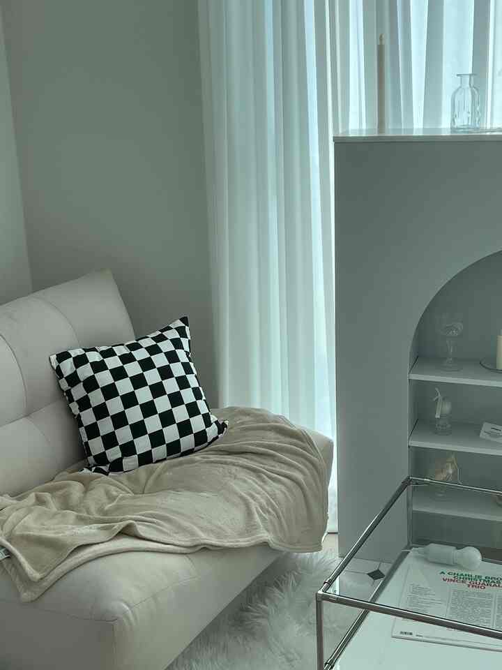 Ivory and beige toned small room featuring a single sofa with checkerboard cushion and blanket, modern fireplace console, and transparent glass table creating a calm clean atmosphere