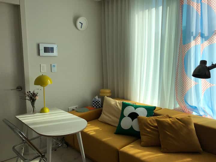 Yellow-toned studio living room featuring a yellow sofa, transparent acrylic chair, white table, and curtains creating a cozy atmosphere