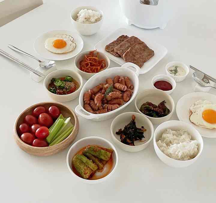 A white-toned dining table set with various Korean side dishes and plates arranged neatly for a two-person meal in a newlywed home