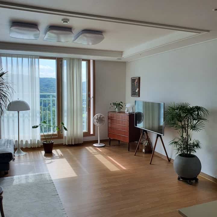 Bright living room with white walls and natural wood floors, featuring a TV stand, plants, and floor lamp in a natural setting