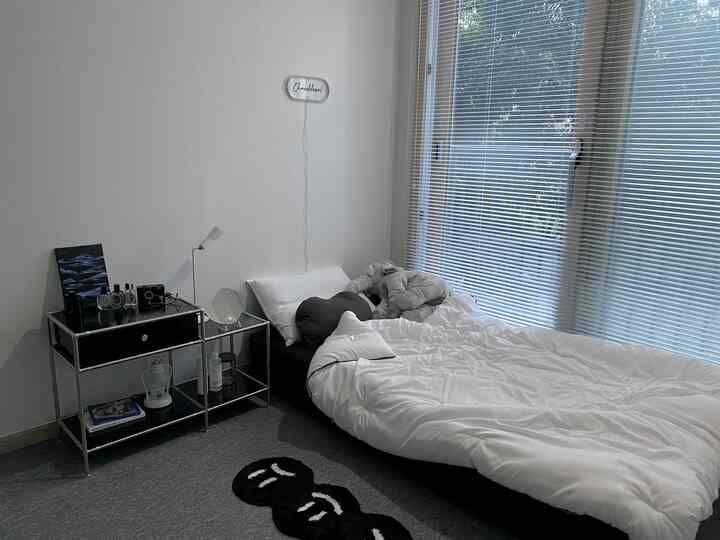 Monotone bedroom with white walls and bedding, black bed frame and nightstand, and large window equipped with aluminum blinds